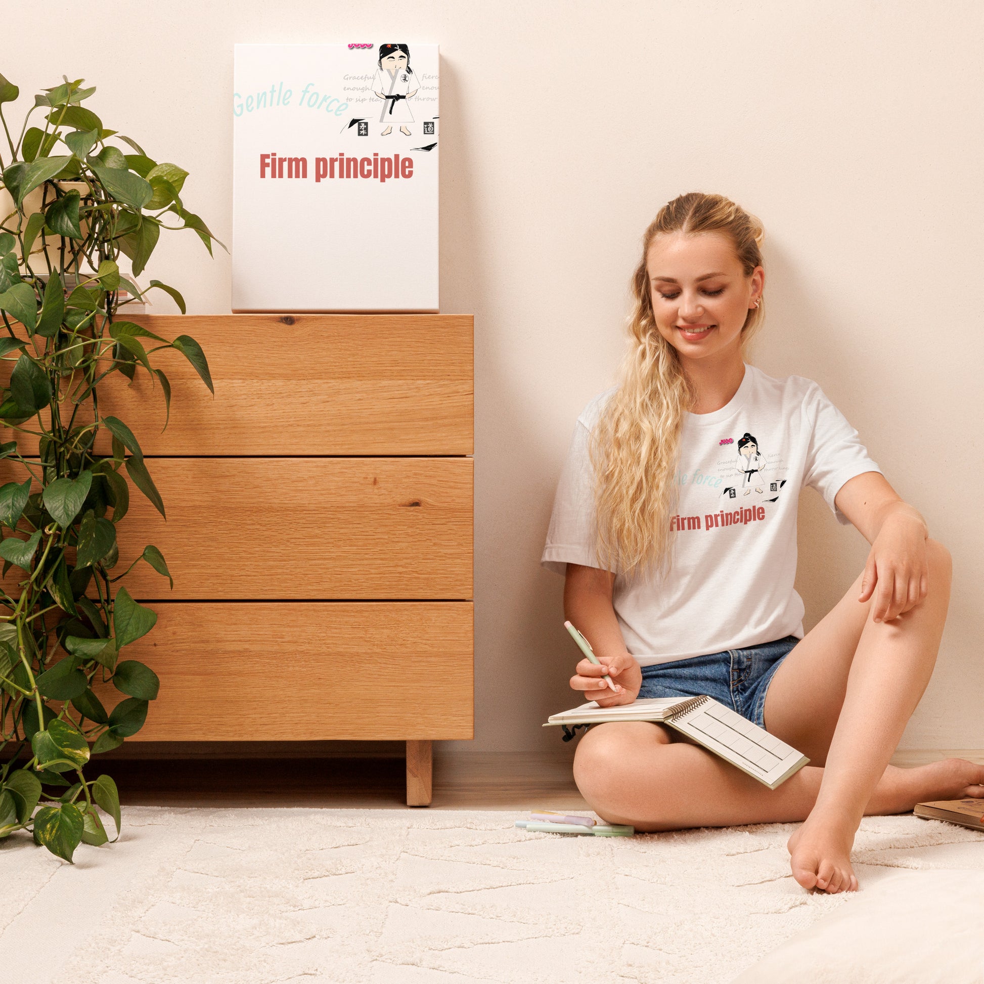 Woman wearing a white tee with a judo lady graphic design and motivational quote sitting on the floor reading a book with a 'Firm Principle' t-shirt and poster in the background.